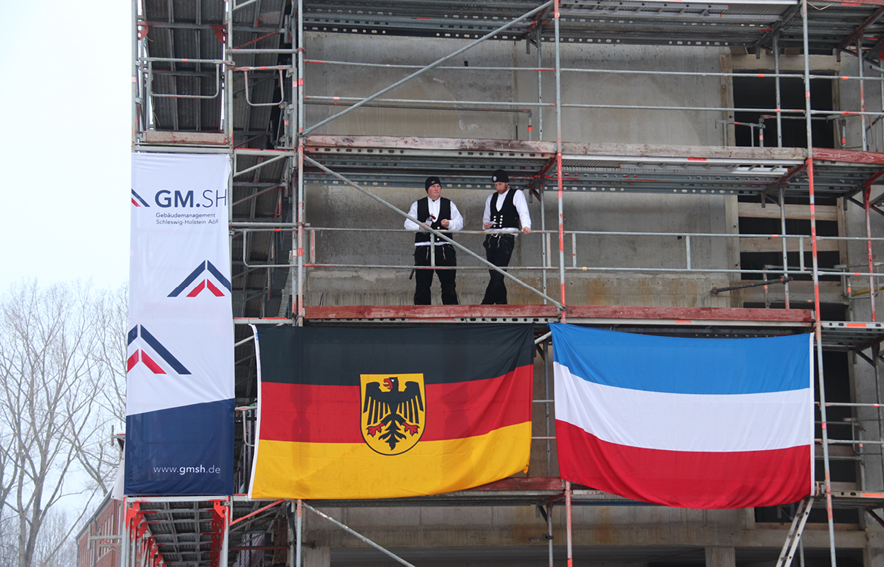 zwei Handwerker auf Gerüst, an dem drei Flaggen hängen: GMSH, Deutschland, Schleswig-Holstein